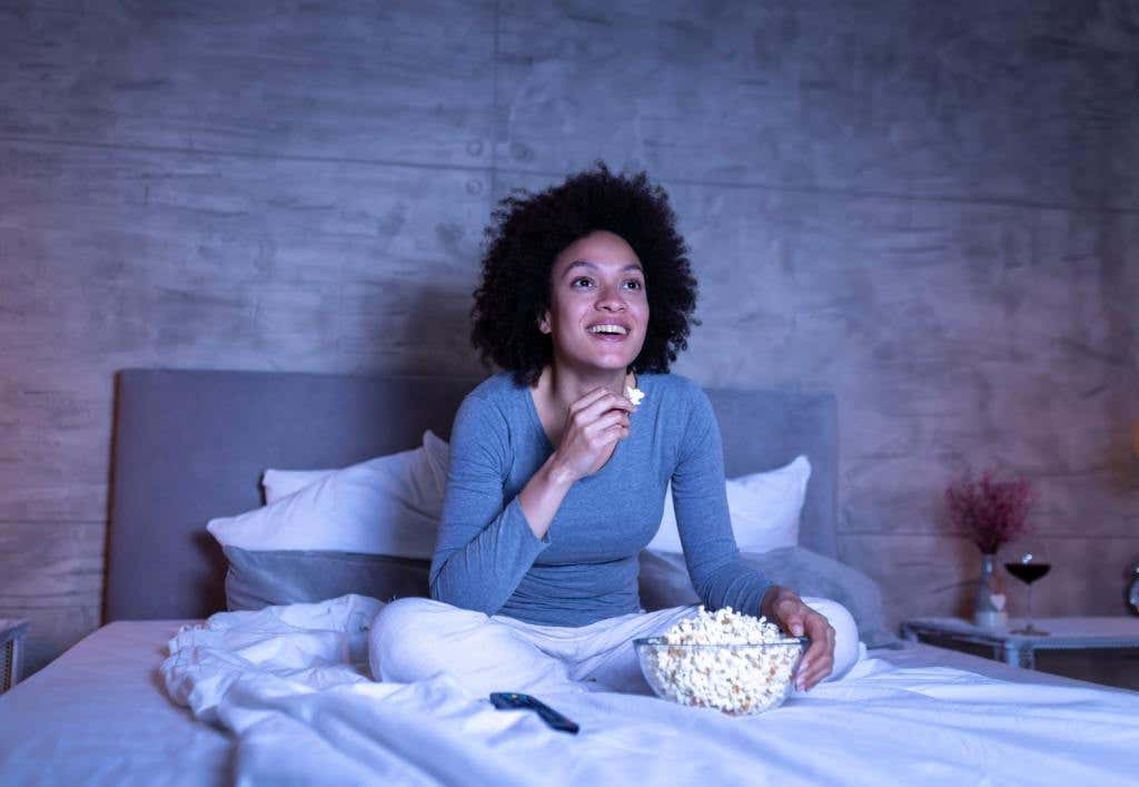 Beautiful mixed race woman wearing pajamas sitting on bed, eating popcorn and watching a comedy movie on TV, relaxing at home late at night