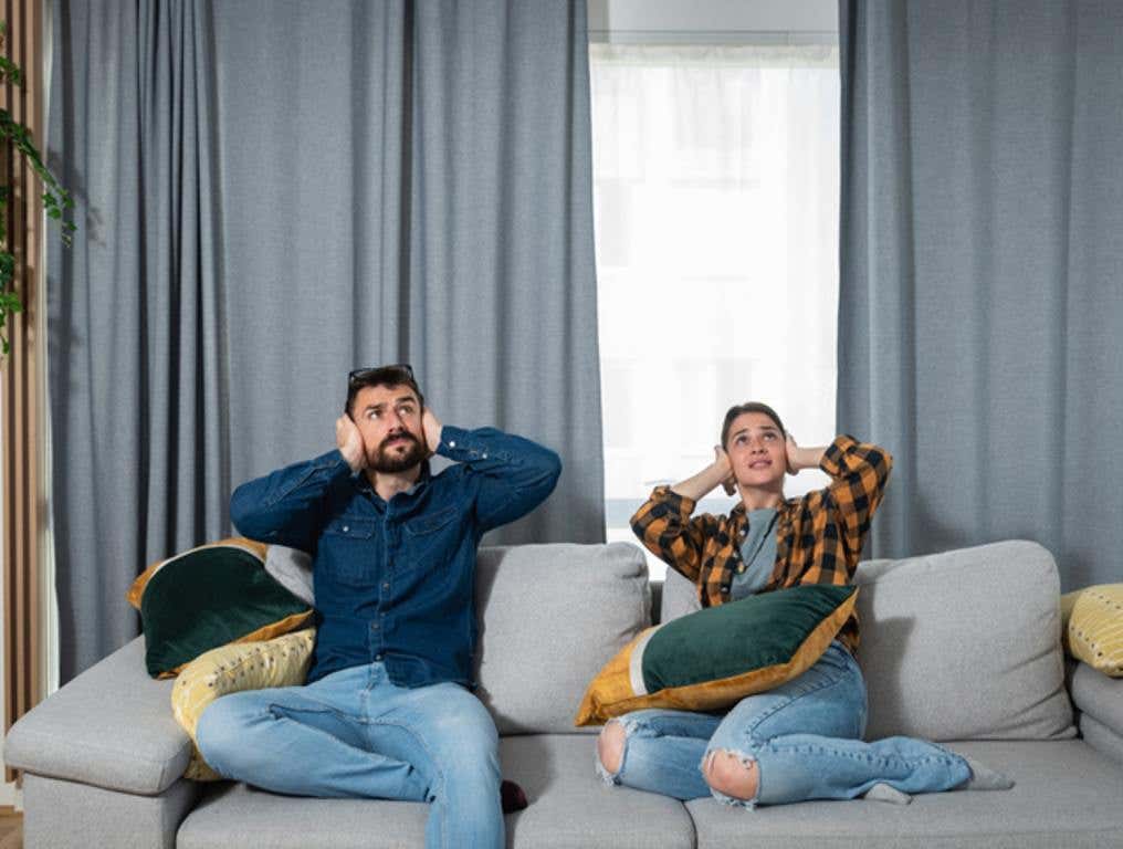 Young couple is sitting on a sofa in their apartment looking up and holding their hands to plug their ears as a neighbor upstairs is having a party and playing loud music or renovating the apartment