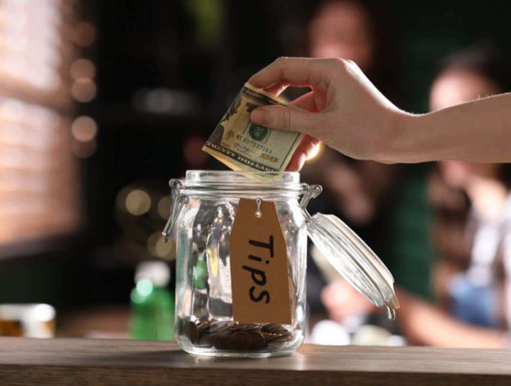 Woman putting tips into glass jar on wooden table indoors, closeup, tipping concept