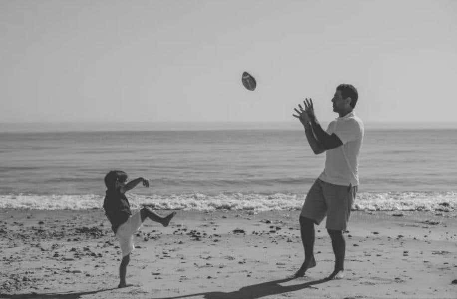 A young kid throws a football to his dad on the beach.