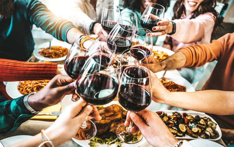 Happy friends toasting red wine glasses at dinner party over a table of food.