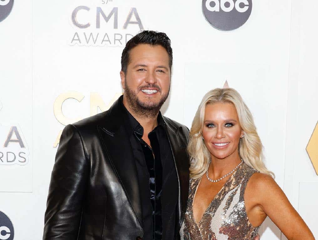 Luke Bryan in a black leather jacket and his wife Caroline in a gold dress on a CMA red carpet