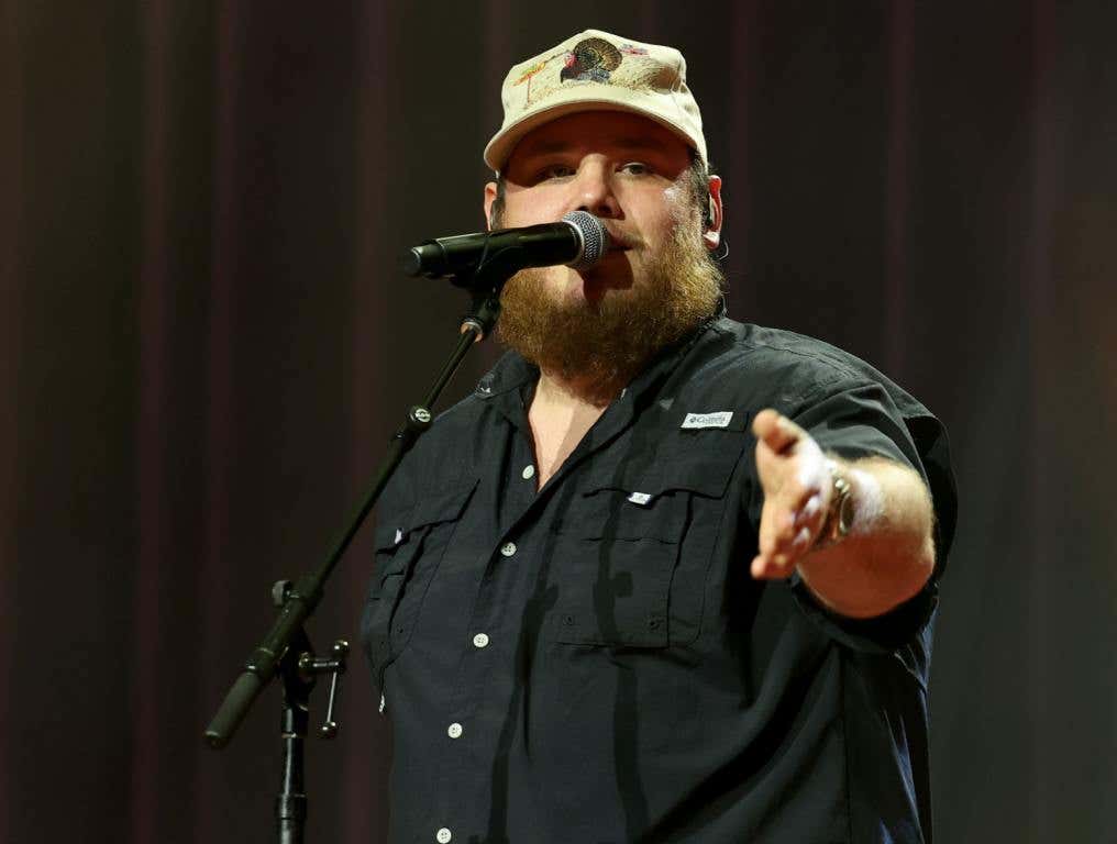 Luke Comsb performing on stage in a black shirt and tan ball cap.