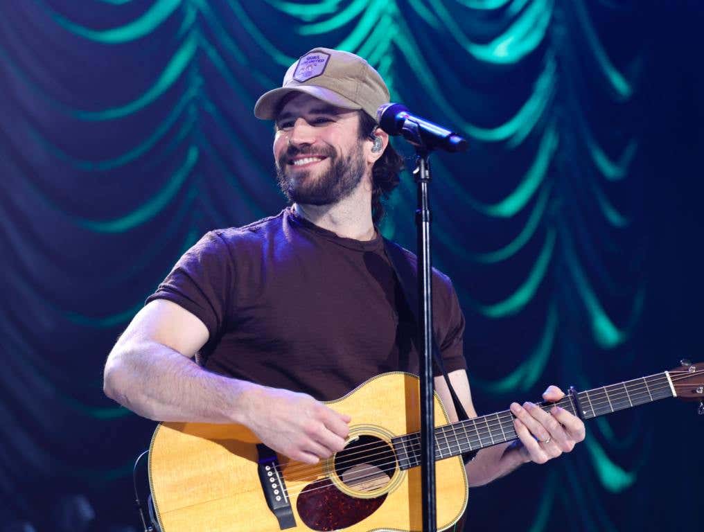 Sam Hut playing guitar on stage in a black shirt and tan ball cap.