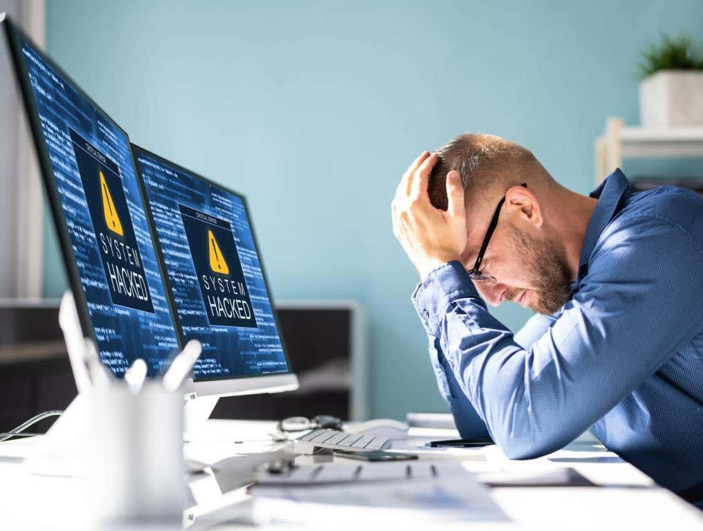 A white man in a blue business shirt is sitting at a white desk with his head in his hands after his two computer screens show that he has been hacked. Concept of online payment scams.