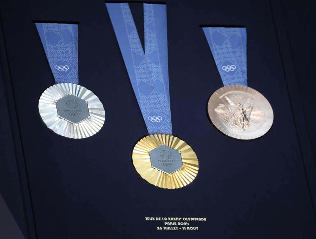 Straight on view of the Paris Olympics Medals for which Boulder City's Lexi Lagan will be shooting. On the left, the silver medal with a blue ribbon. The gold medal sits lower against the black velvet background in the middle of the frame. The bronze medal is on the right. All have blue ribbons attached to the medals. The inscription on the front of each medal reads "Paris 2024" with the five Olympic rings underneath.