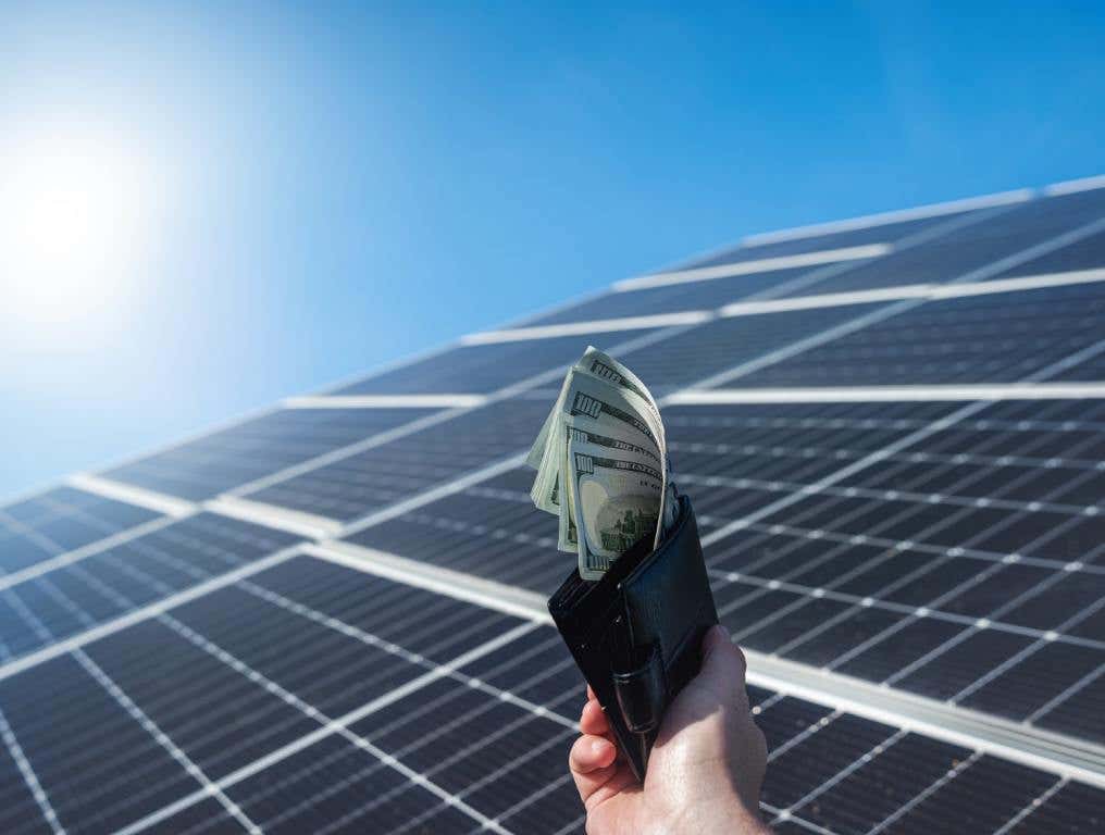 Deceptive solar companies have led to over 1,000 annual complaints to the Nevada State Contractors Board. Photo of a man's right hand holding up a black leather wallet filled with American one hundred dollar bills. The bills are pulled out of the wallet and fanned. Behind the man's hand and wallet are several black and silver newly installed solar panels. Behind that, a bright blue sky and hazy sunshine can be seen.