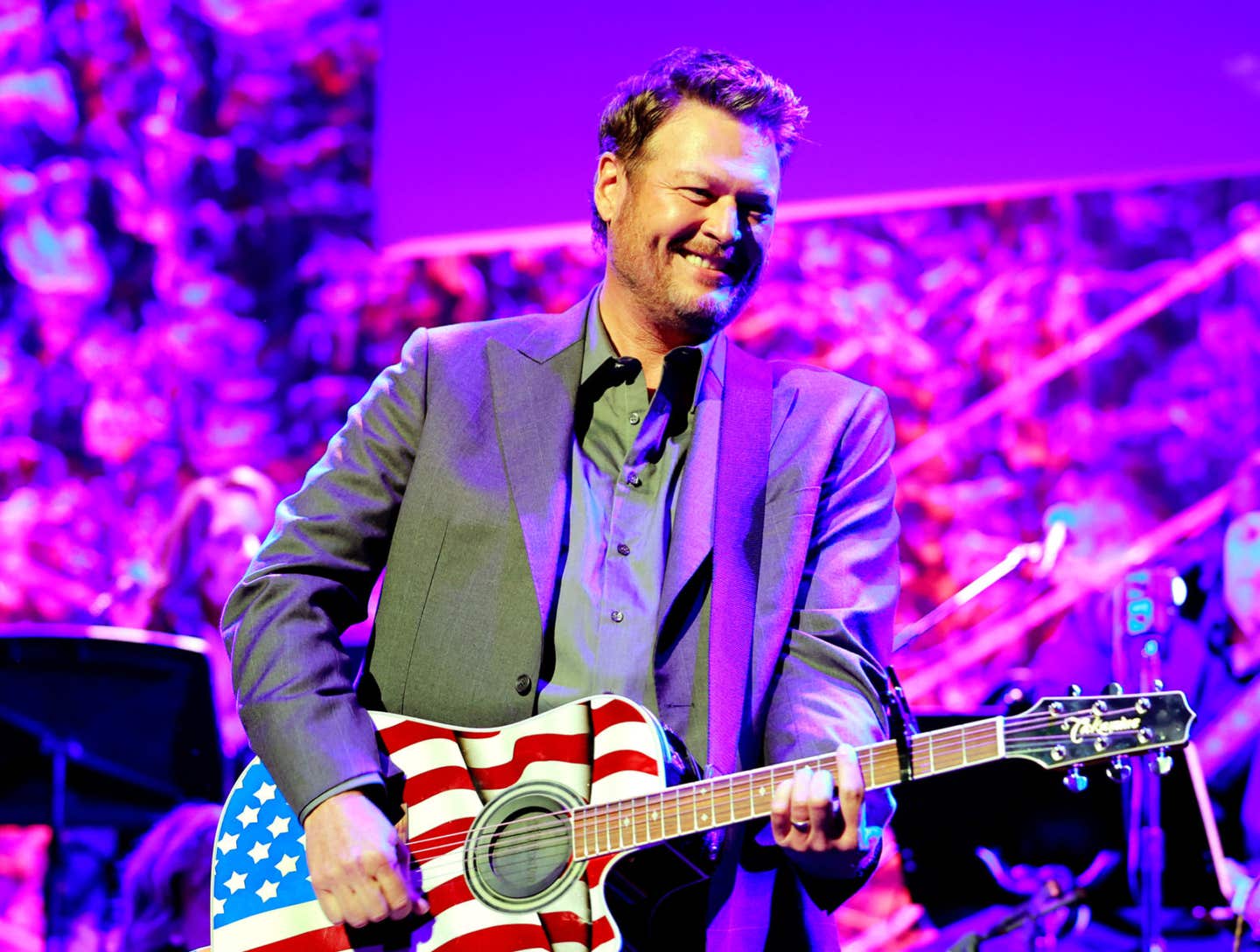 Blake Shelton is on stage playing an American flag guitar in a brown blazer.