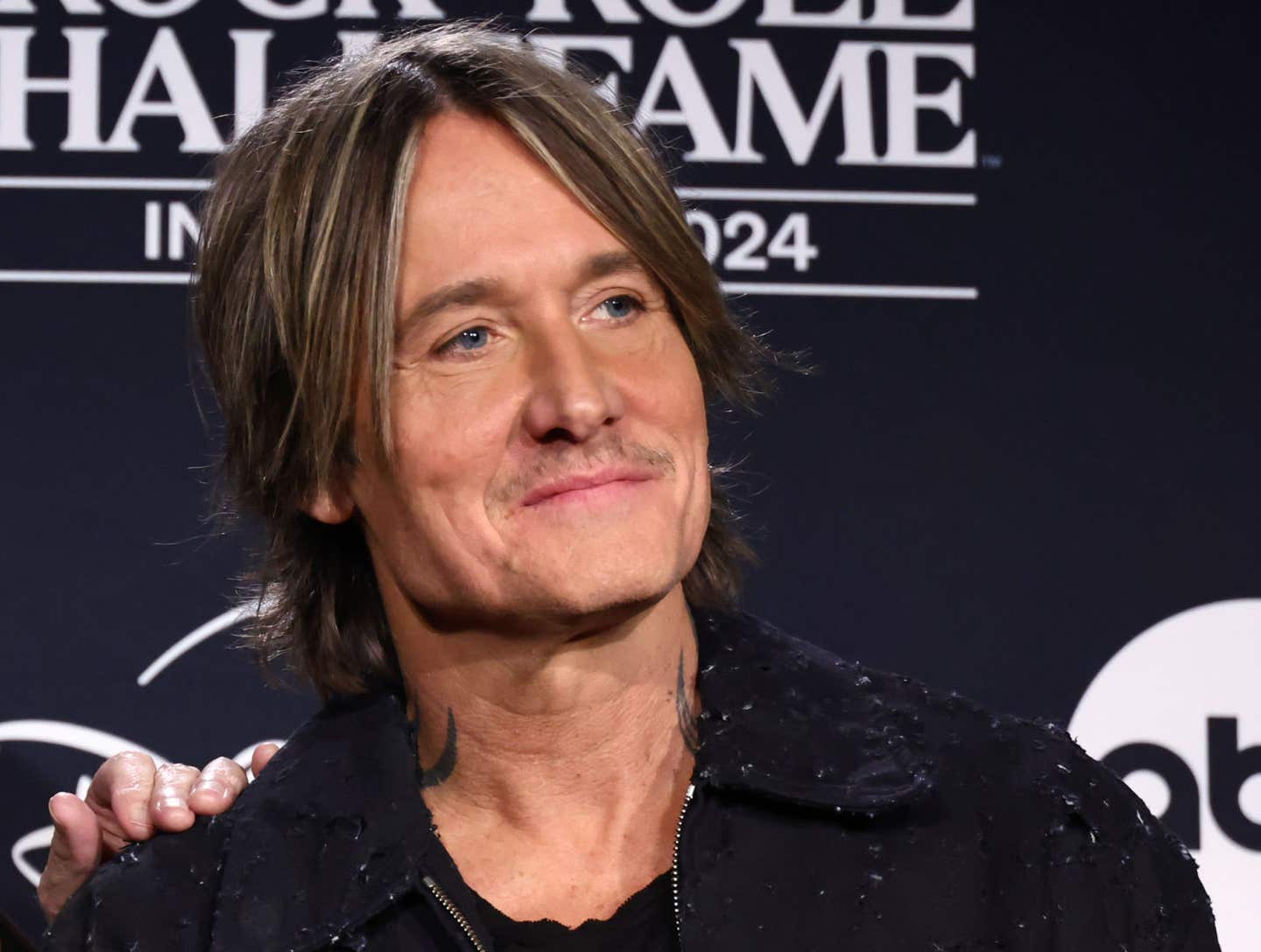 Keith Urban poses on a red carpet wearing a black shirt and a black denim jacket.