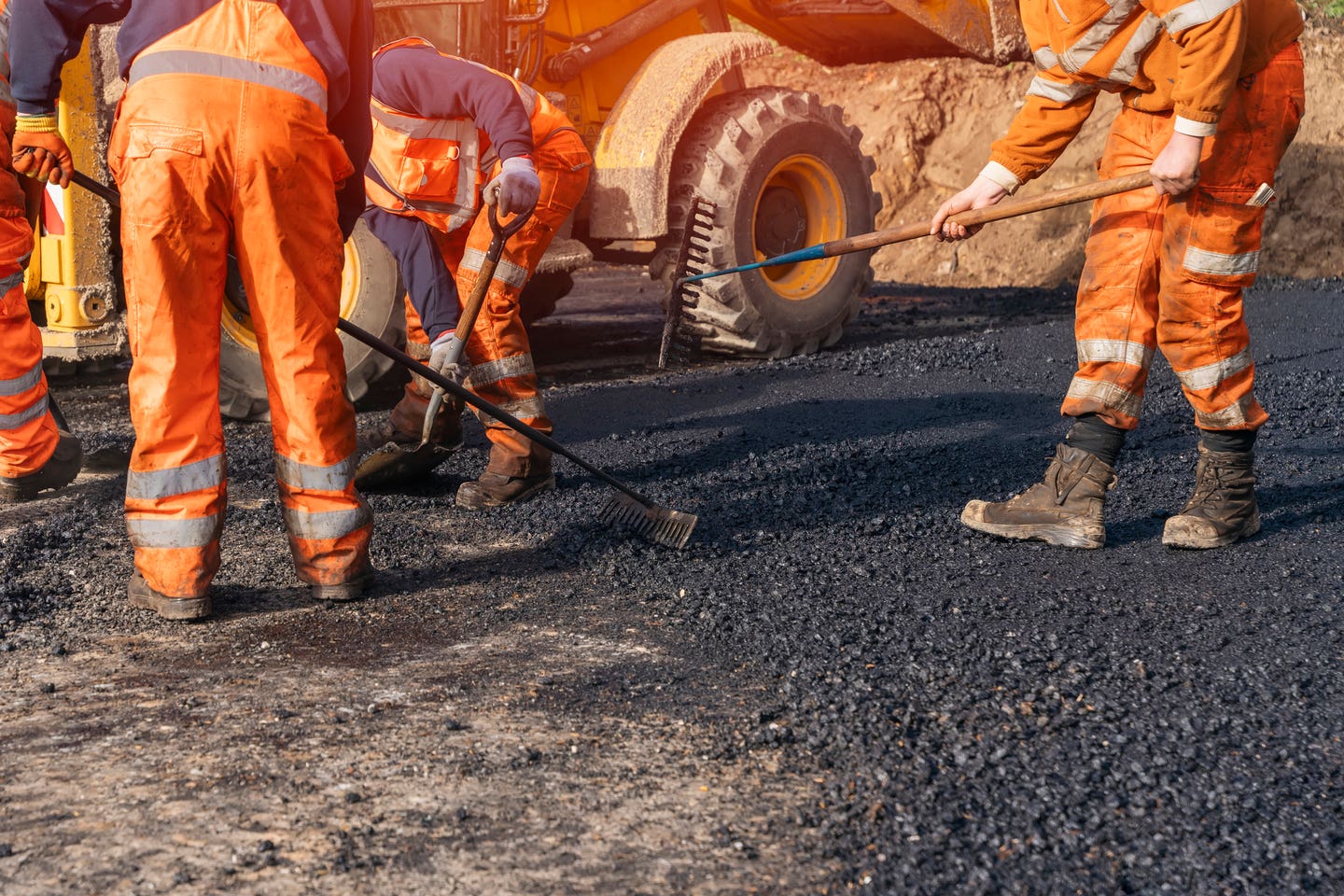 A group of construction workers in bright orange uniforms diligently lay down asphalt on a roadway. Heavy machinery is visible in the background, adding to the busy atmosphere of the worksite.