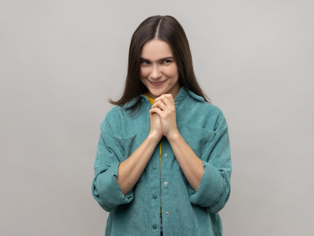 Devious cunning young woman with dark hair clasping hands and smirking mysteriously, scheming cheats, evil prank, wearing casual style jacket. Indoor studio shot isolated on gray background.