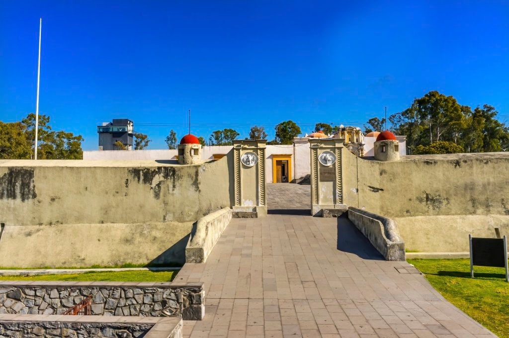 A cobblestone walkway leads up to a a short stone wall with an opening. Behind it is a small stucco building with a flat roof and mustard yellow door.