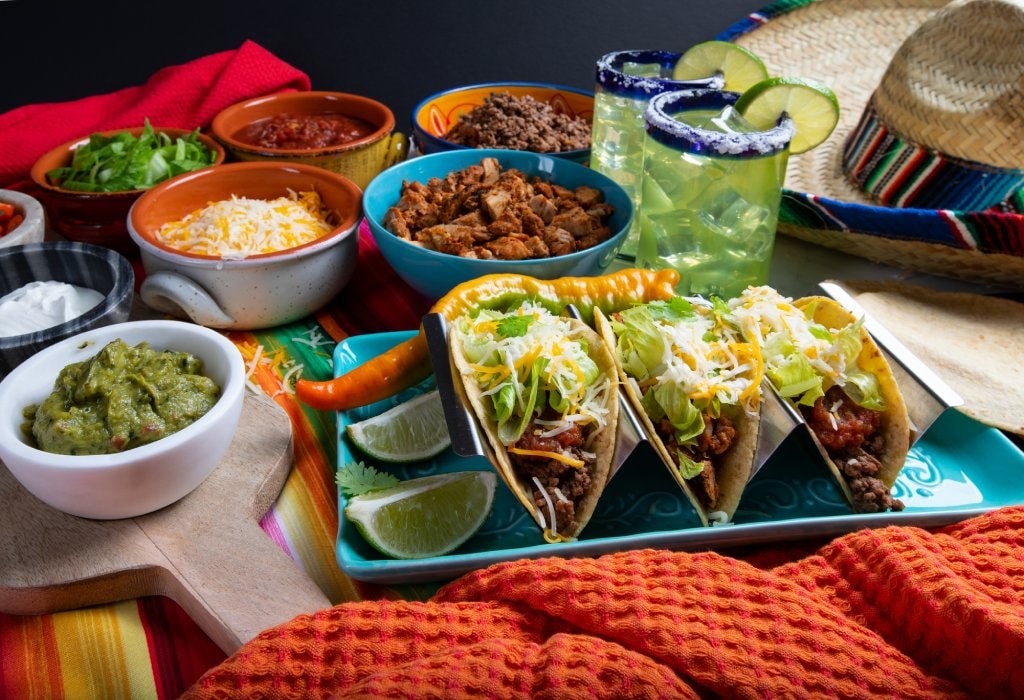 Tacos on a colorful plate with all the trimmings like chicken, beef, sour cream and cheese, in bowls surrounding. The spread is on a rustic background to be served in a restaurant with sombrero with margaritas.