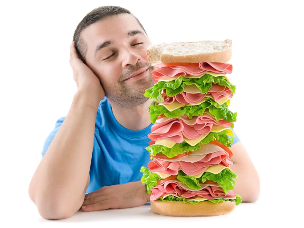 A man in a blue t-shirt sits at a white table with his head in his hand and a content smile on his face. Next to him is a very tall "Dagwood" sandwich layered with meats, cheeses and lettuce.