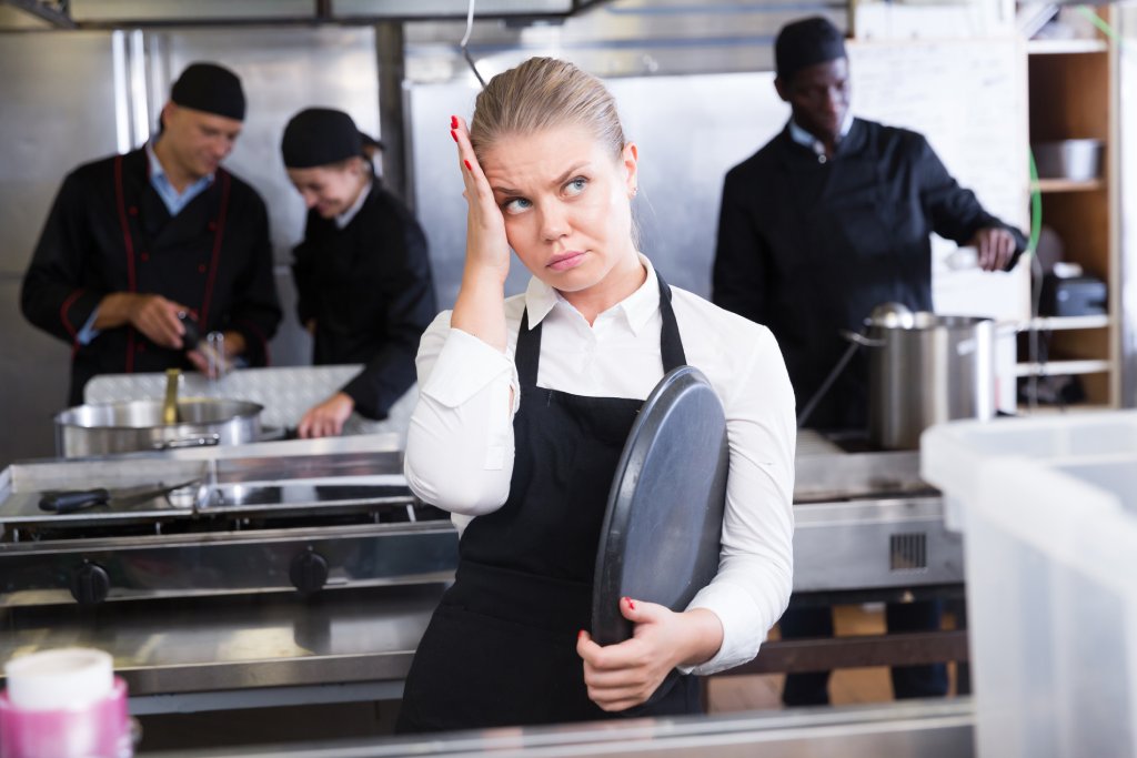 A young blonde girl in a white shirt and black apron is holding a tray in a commercial kitchen as she looks disappointed with her head in her hand.