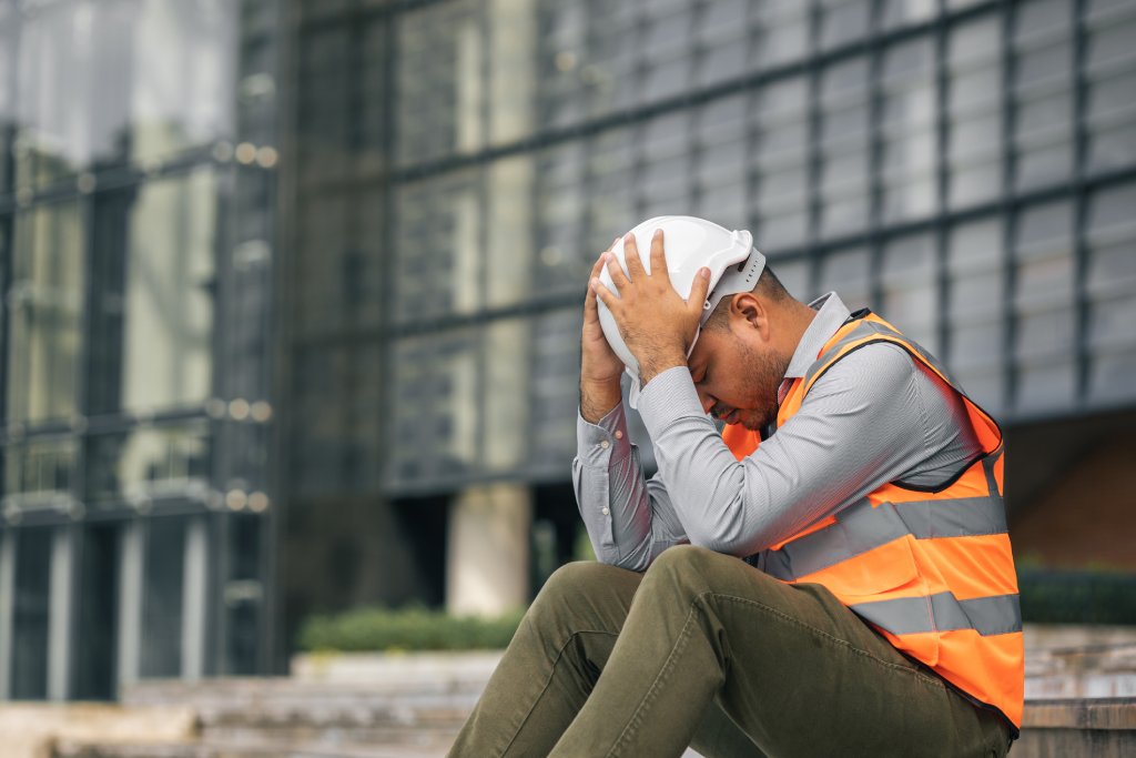 A male construction worker in a vest and hard hat sits on a curb with his head in his hands.