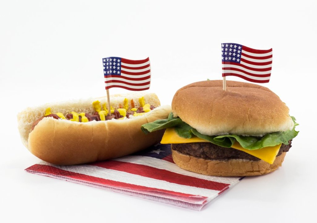 A hot dog and hamburger sit on a white table atop an American flag napkin with American flag toothpicks stuck in them.