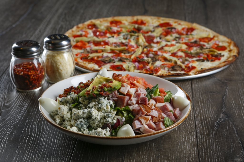 A chopped salad in a shallow white bowl sits in front of a whole pizza on a table. There are parmesan and red pepper shakers to the left.