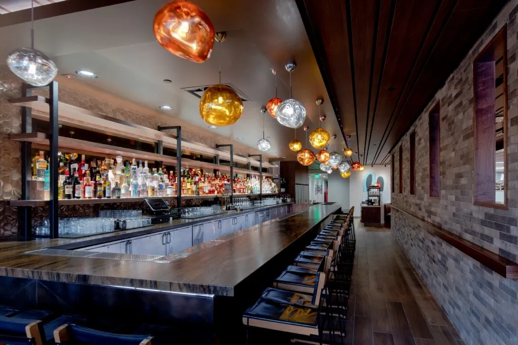 The interior of a bar with brick walls, a dark brown wooden bar, low back bar stools and blown glass light fixtures.